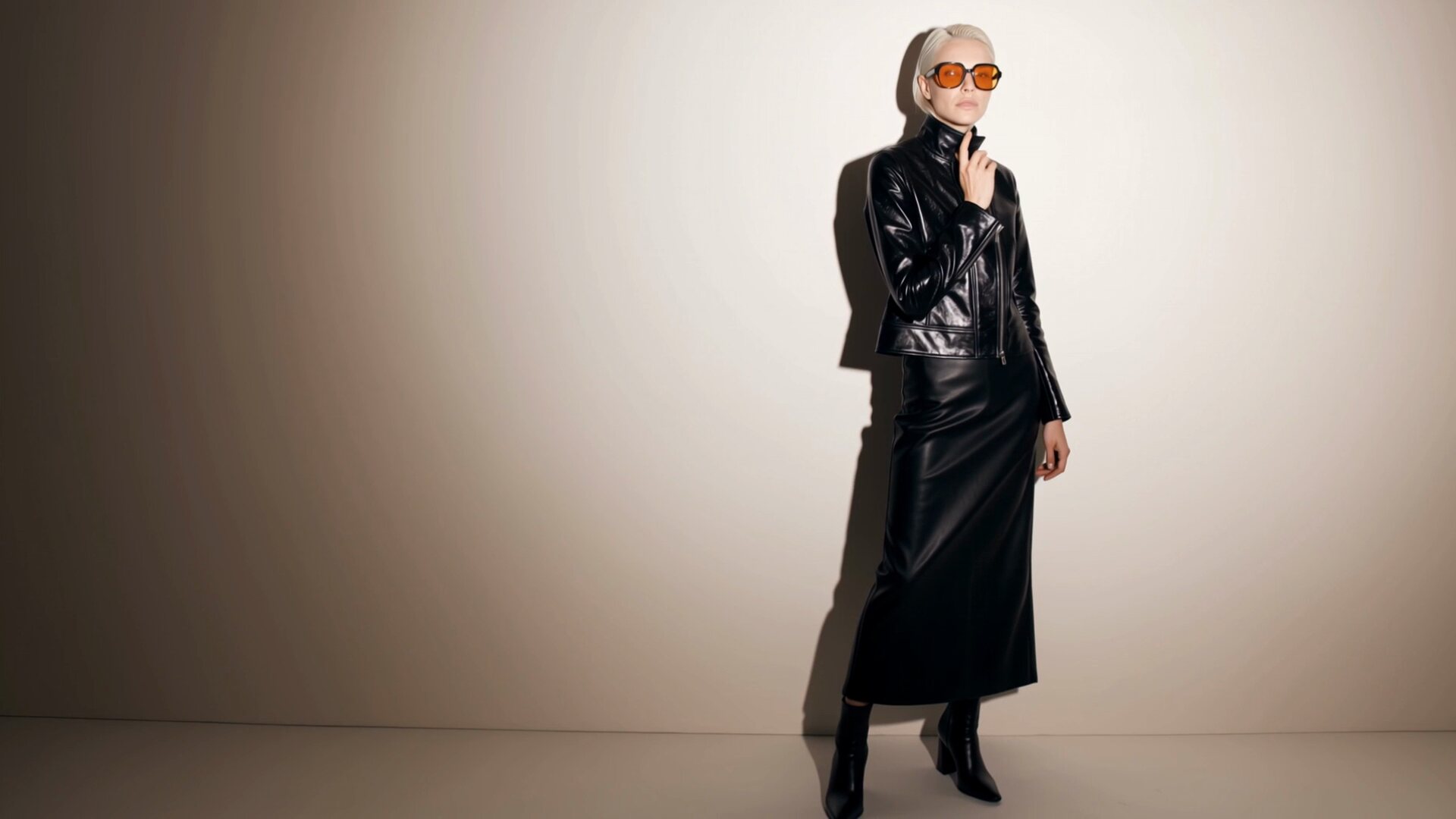 A platinum blonde model in a glossy black leather jacket, long leather skirt and orange tinted sunglasses stands against a white studio wall.